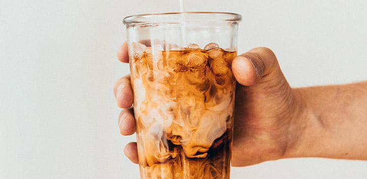 A hand holding a clear glass with milk being poured into the coffee