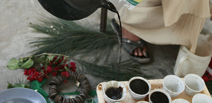 A woman pours coffee from a black kettle into a line of small, handle-less cups on a tray