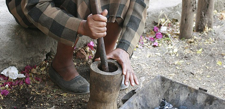 A person crouches on the ground and grinds coffee beans in a mortar and pestle