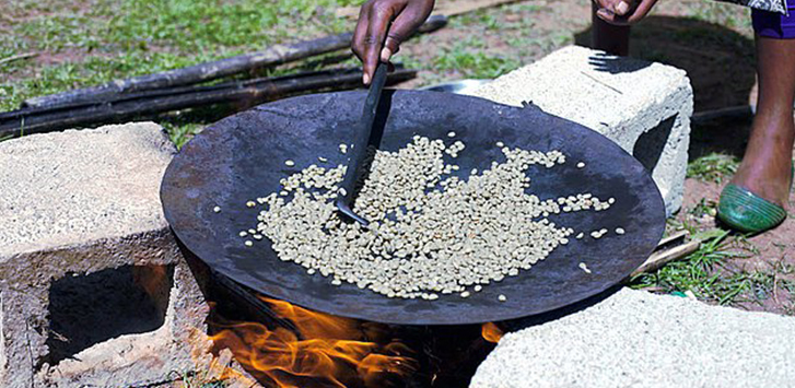 A person stirs coffee beans in a black bowl over a fire pit