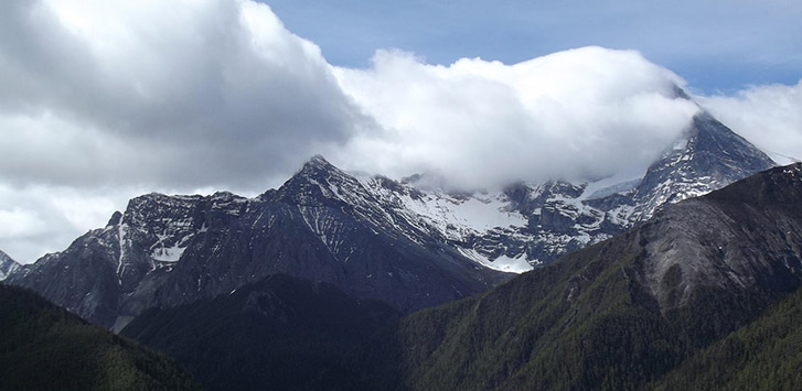 A landscape photo of rocky mountains coated in snow at the top with large white clouds in the sky