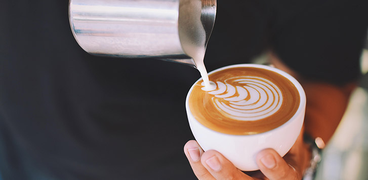 A person pours milk into a white mug, creating a tulip design in the espresso